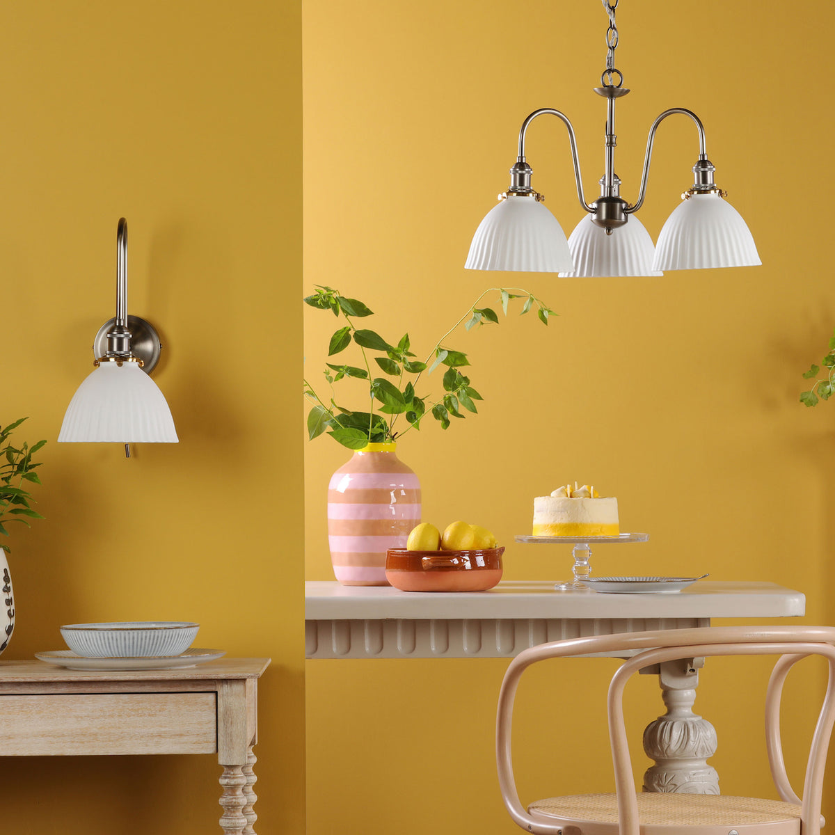 A yellow room with a wall light and a chandelier, a vase of greenery, lemons in a bowl, and a cake.