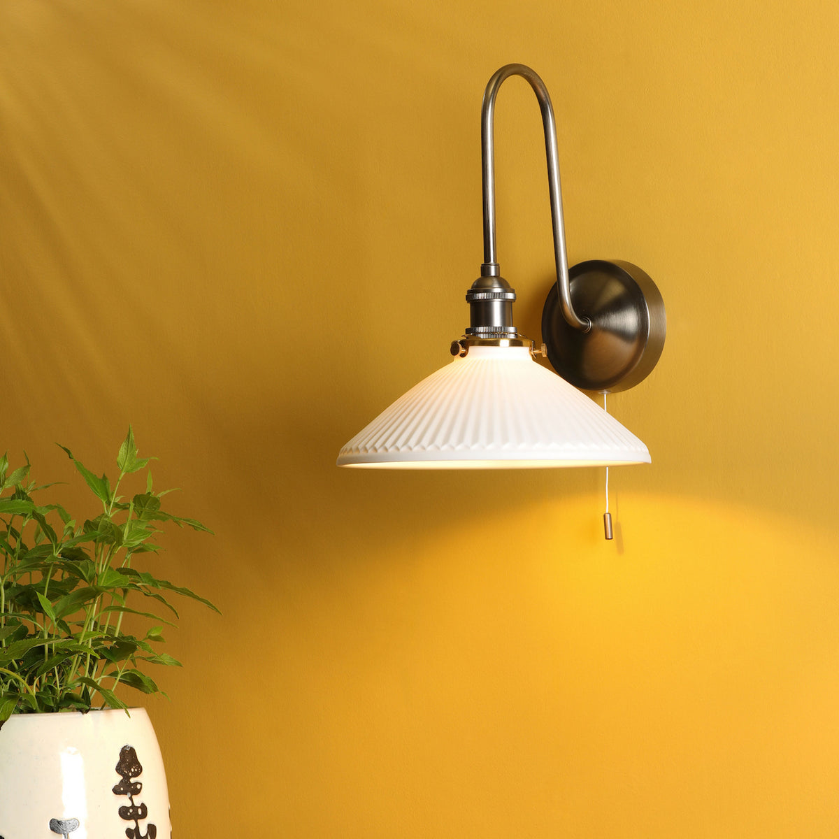 A white fluted wall sconce with a brushed nickel arm and base, illuminating a mustard yellow wall.