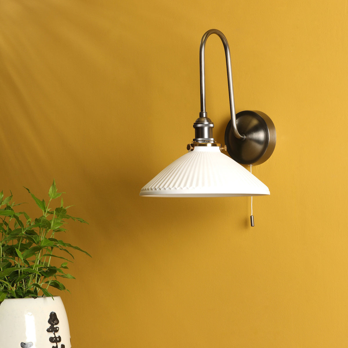 A white fluted wall sconce with a brushed nickel arm and pull chain, mounted on a mustard yellow wall.