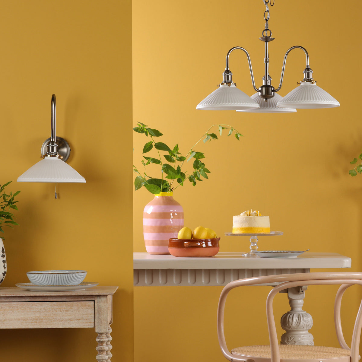 A dining room with yellow walls, featuring a wall light and a three-arm chandelier with pleated shades.