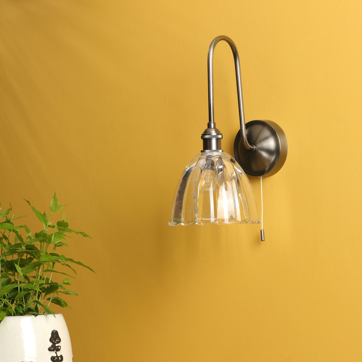 A wall-mounted light fixture with a clear, fluted glass shade and a brushed metal arm on a yellow wall. A green plant in a white pot is in the foreground.