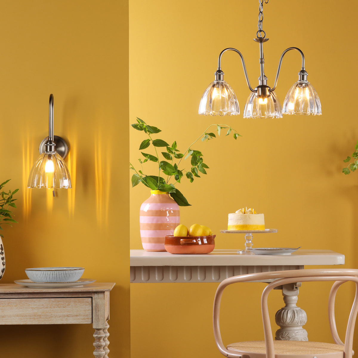 A dining room with yellow walls, a wall light, a chandelier, and a table with a cake and lemons.
