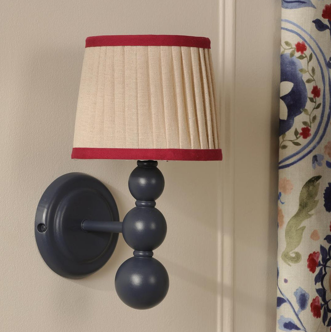 Wall sconce with a navy blue stacked ball base and a pleated beige lampshade with red trim