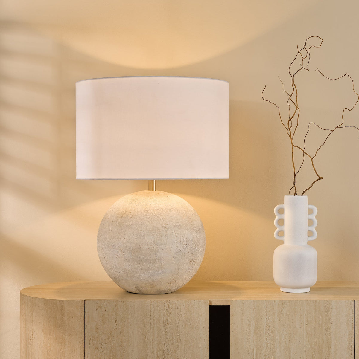 Table lamp with a round base and a white lampshade next to a white vase with dried branches