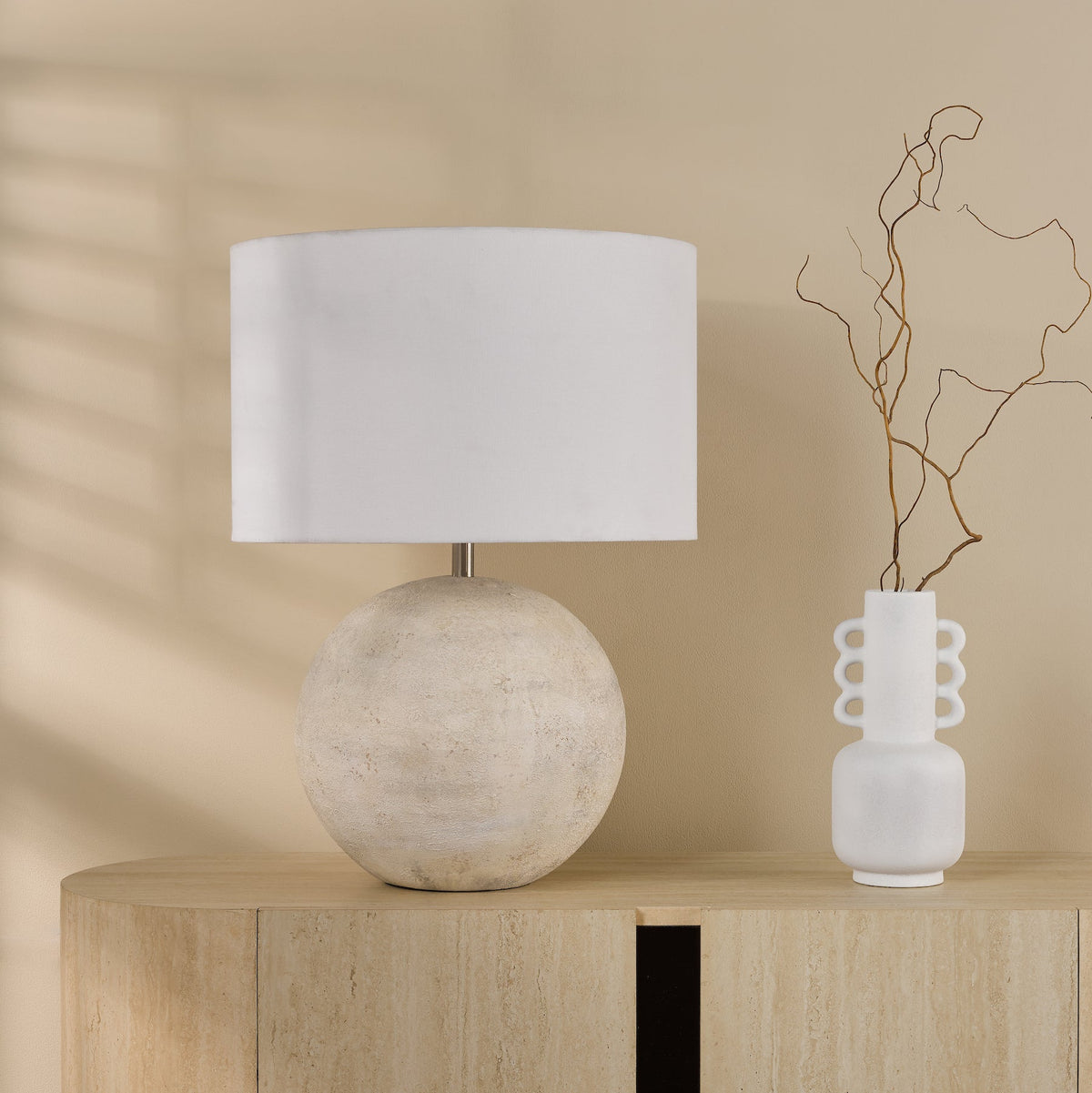 Table lamp with a round textured base and a white cylindrical shade, next to a white vase with dried branches