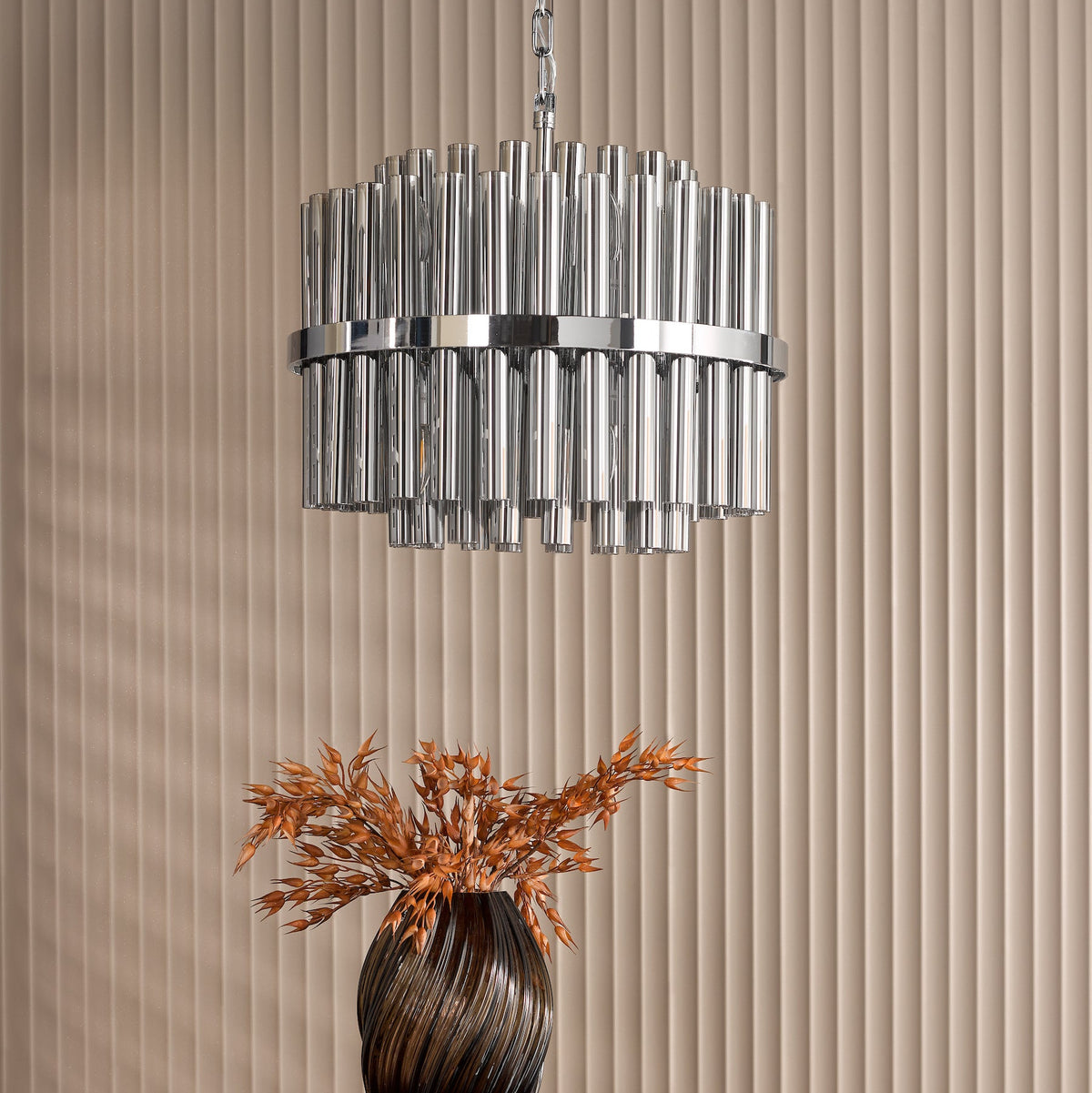 Chrome chandelier with glass rods and a brown vase with dried wheat on a beige fluted wall