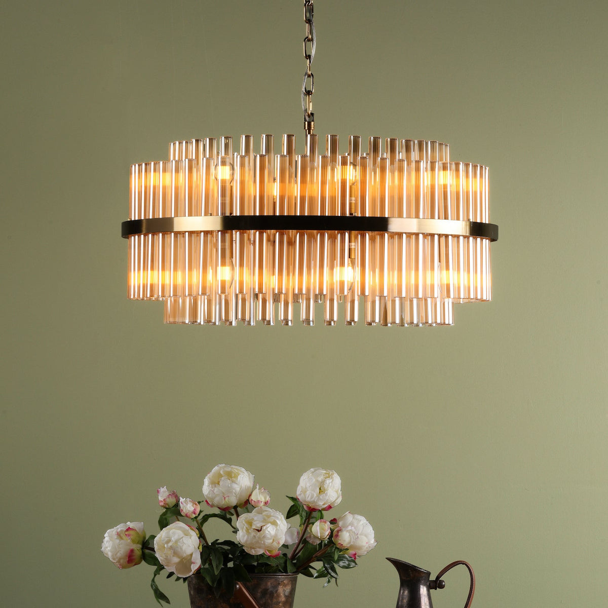 A tiered chandelier with amber glass rods and a gold frame, hanging above a vase of white peonies
