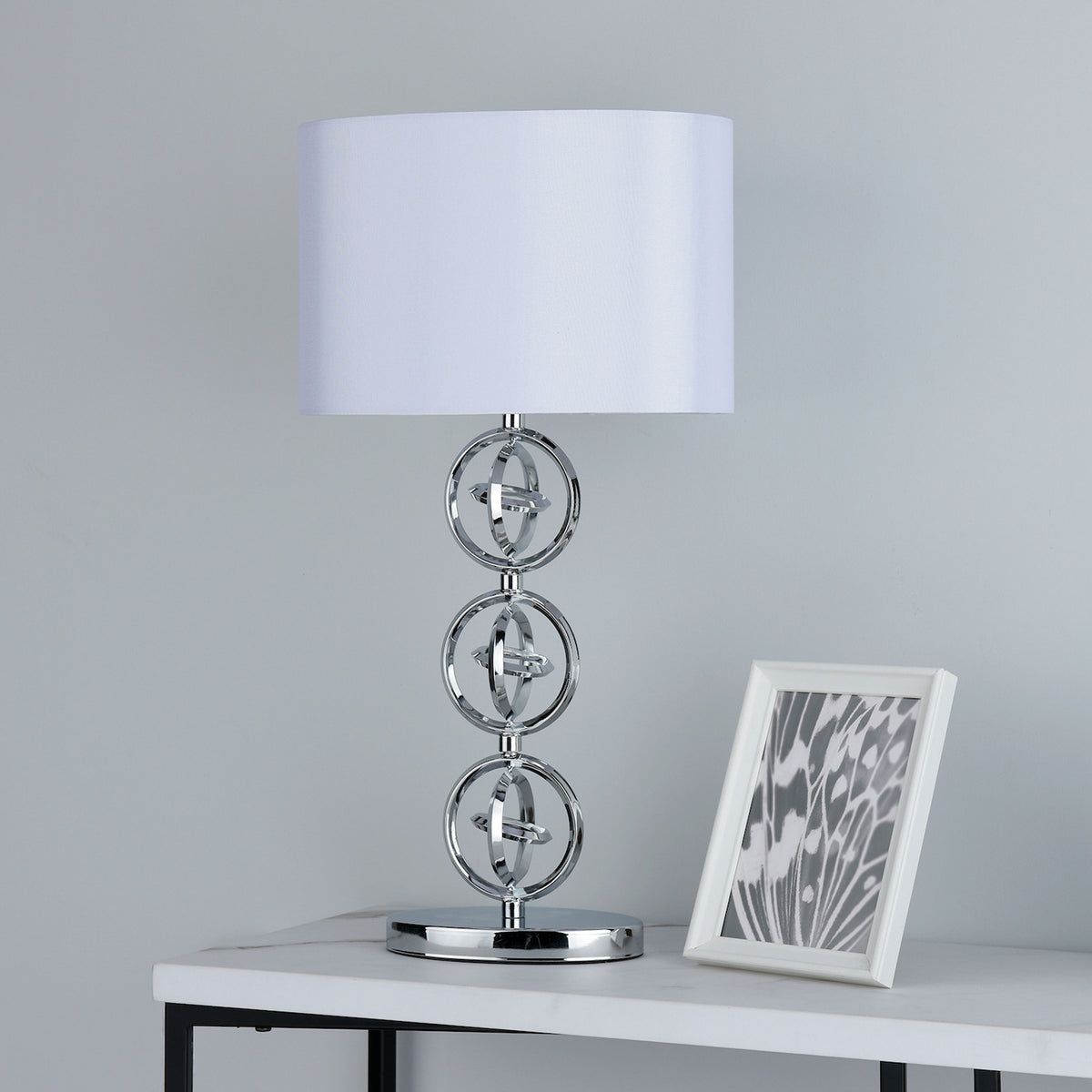 A chrome table lamp with a white shade and a white framed picture on a white marble table.