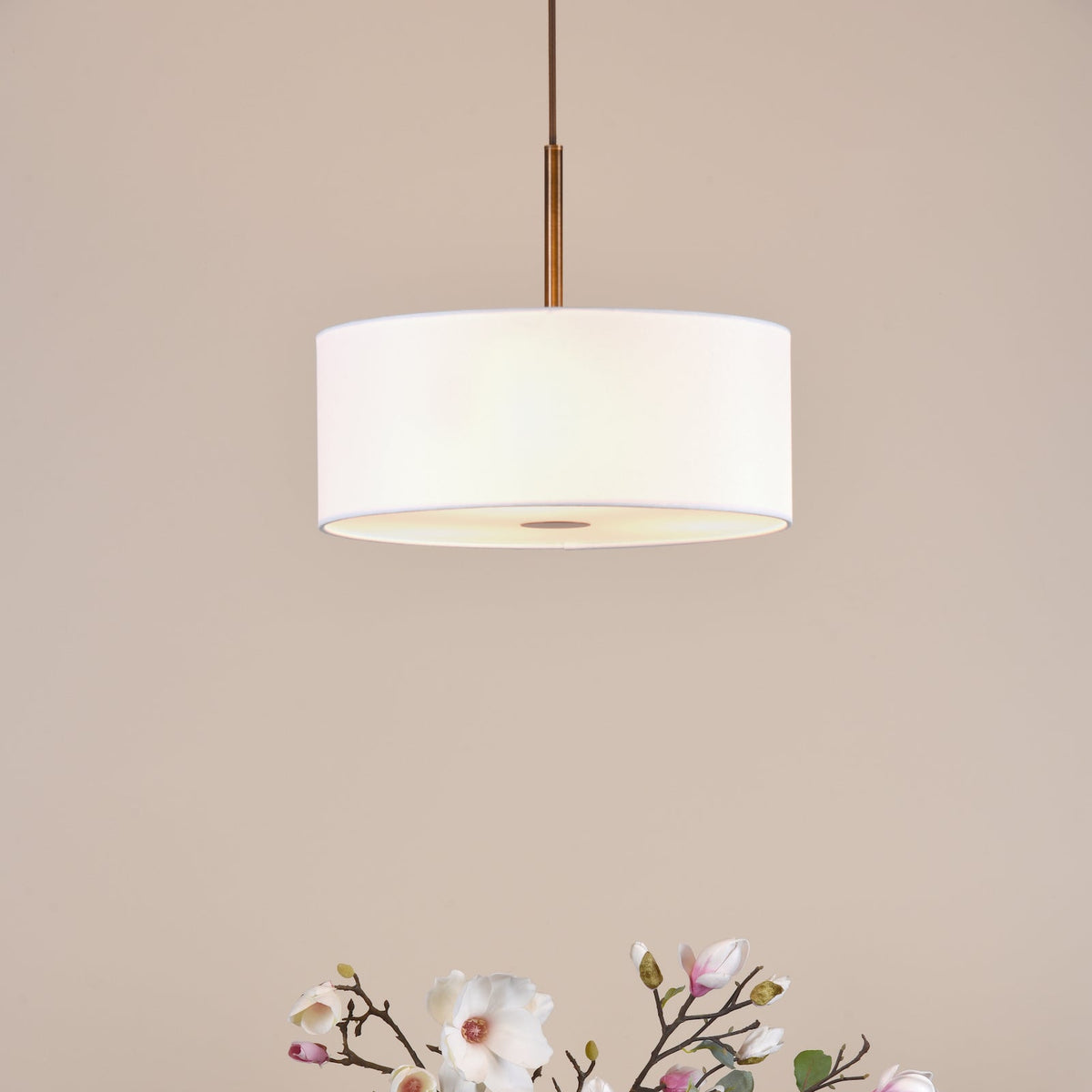 White drum pendant light with a brass rod and brown cord, above magnolia branches with white and pink flowers