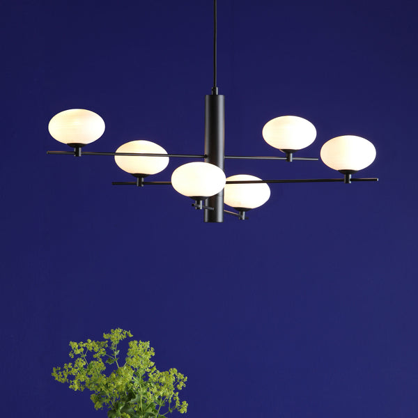 Modern black chandelier with six white globe lights against a deep blue wall, with green foliage in the foreground.