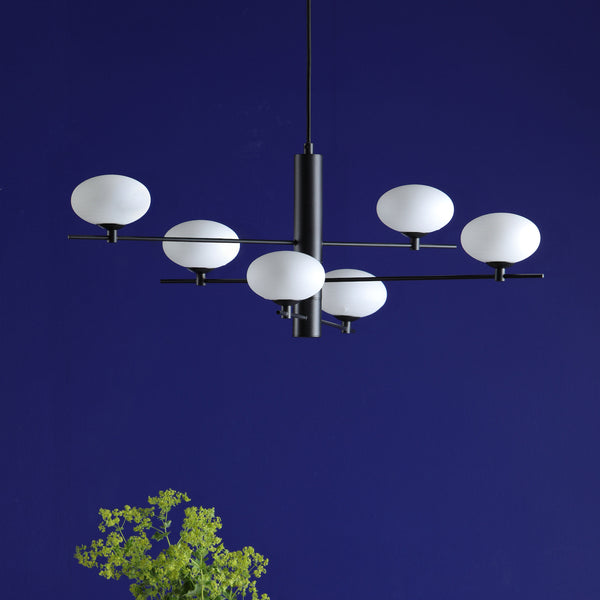 Modern black chandelier with six white globe lights against a deep blue wall, with green foliage below.