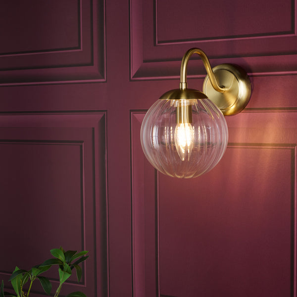 Brass wall sconce with a clear ribbed glass globe shade and exposed bulb on a burgundy wall