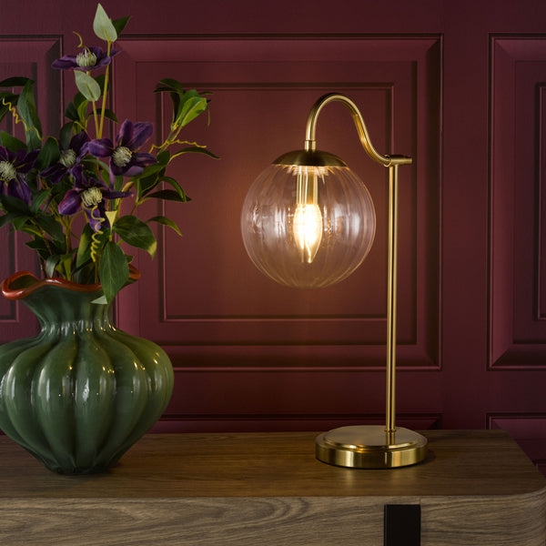 Gold table lamp with a clear glass globe shade and a green vase with purple flowers