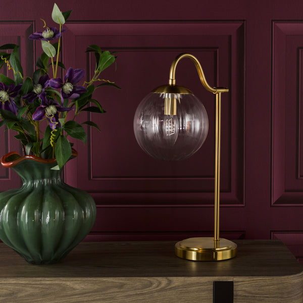 Green vase with purple flowers next to a gold table lamp with a clear glass globe shade