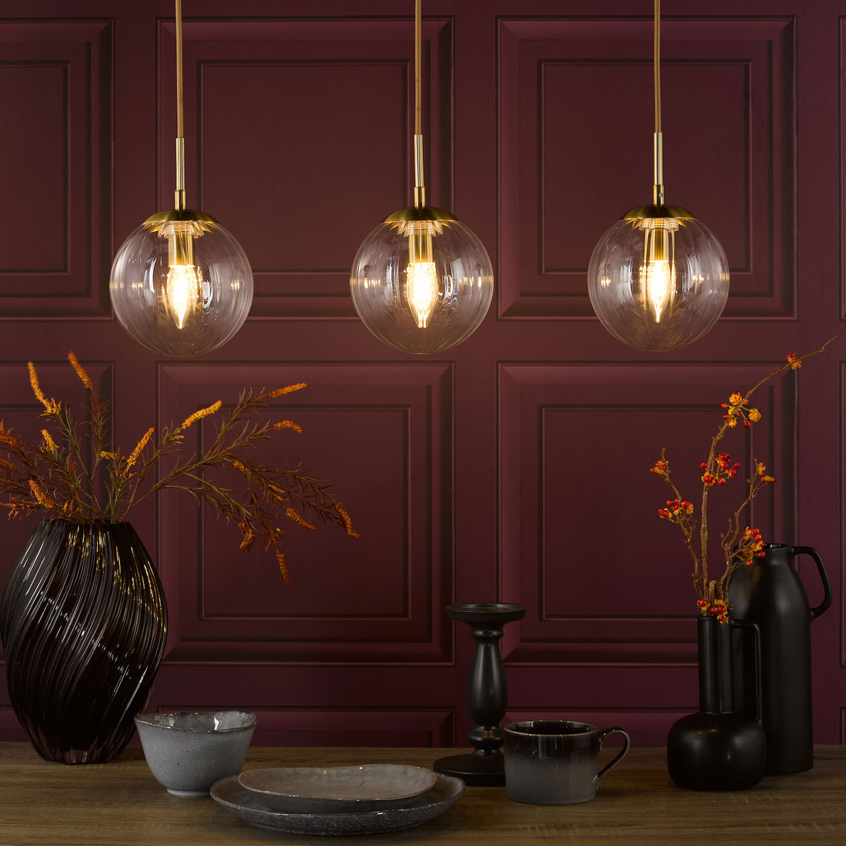 Three glass pendant lights with brass accents illuminate a table set with dark stoneware and vases