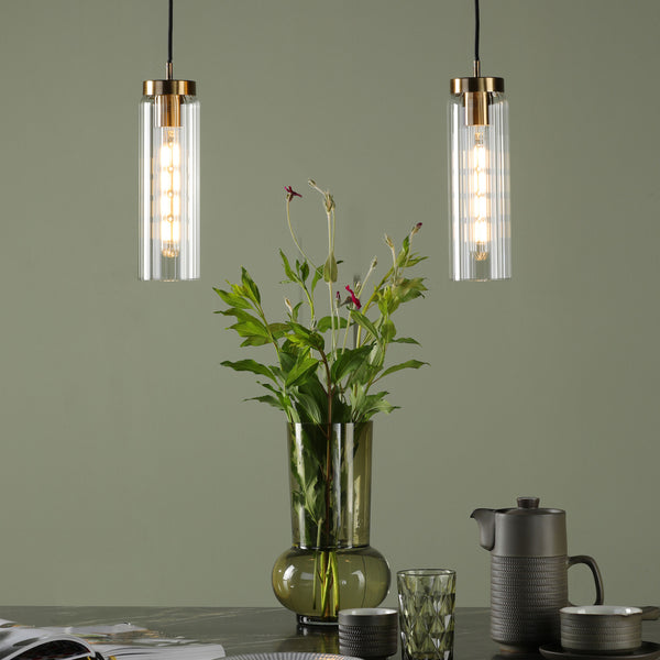 Two pendant lights with ribbed glass shades and brass accents illuminate a vase of greenery and dark stoneware.