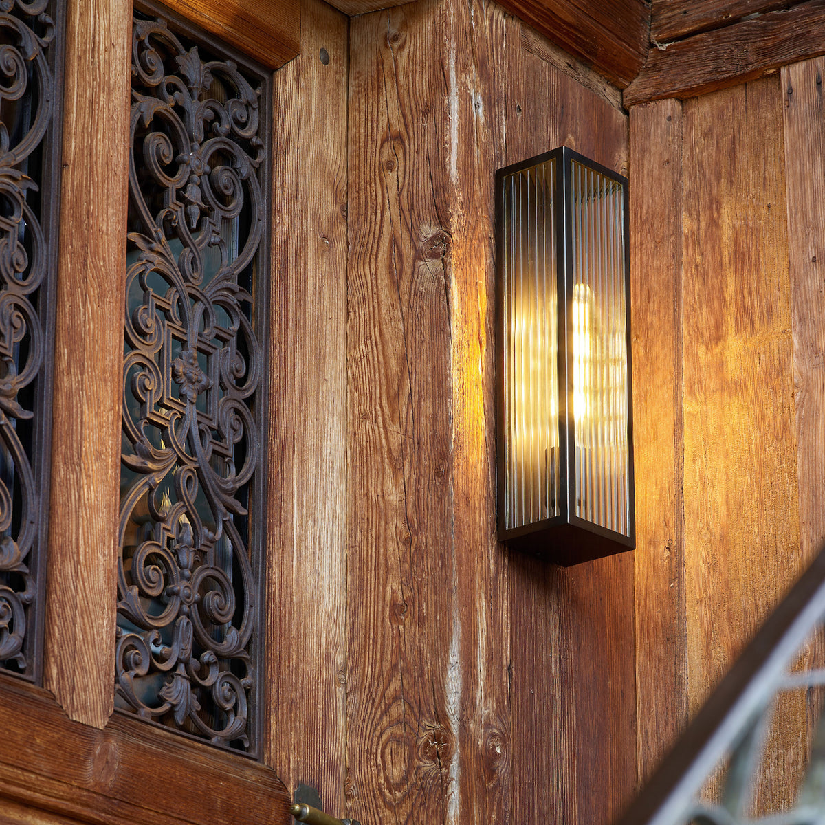 A close-up of a wooden wall with a decorative metal grate and a lit rectangular wall sconce