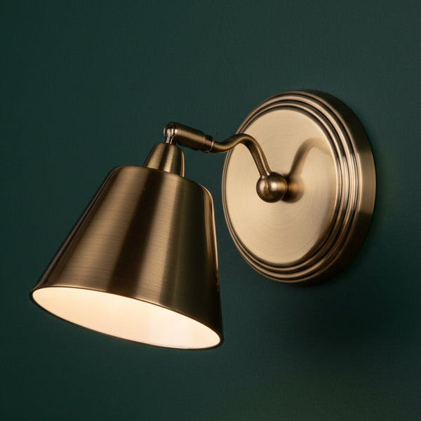 Brass wall light with a conical shade and a round base, mounted on a dark green wall