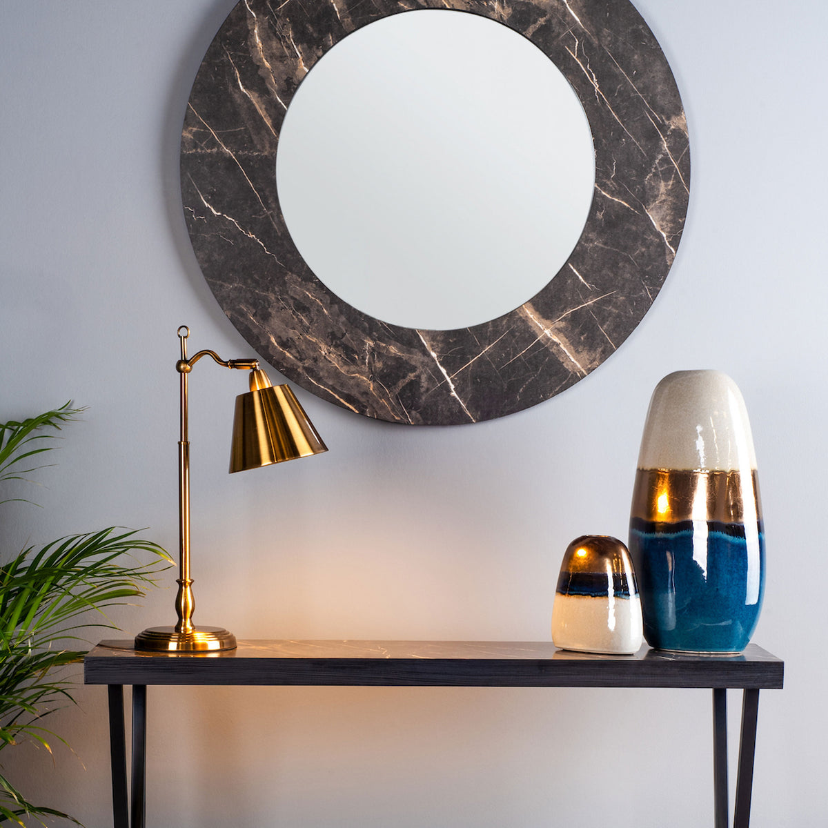 Round mirror with a dark marble frame above a console table with a brass lamp and two decorative vases