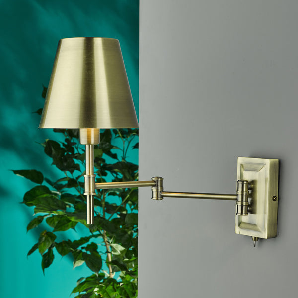 Brass swing arm wall lamp with a conical shade, mounted on a grey wall with a green plant in the background