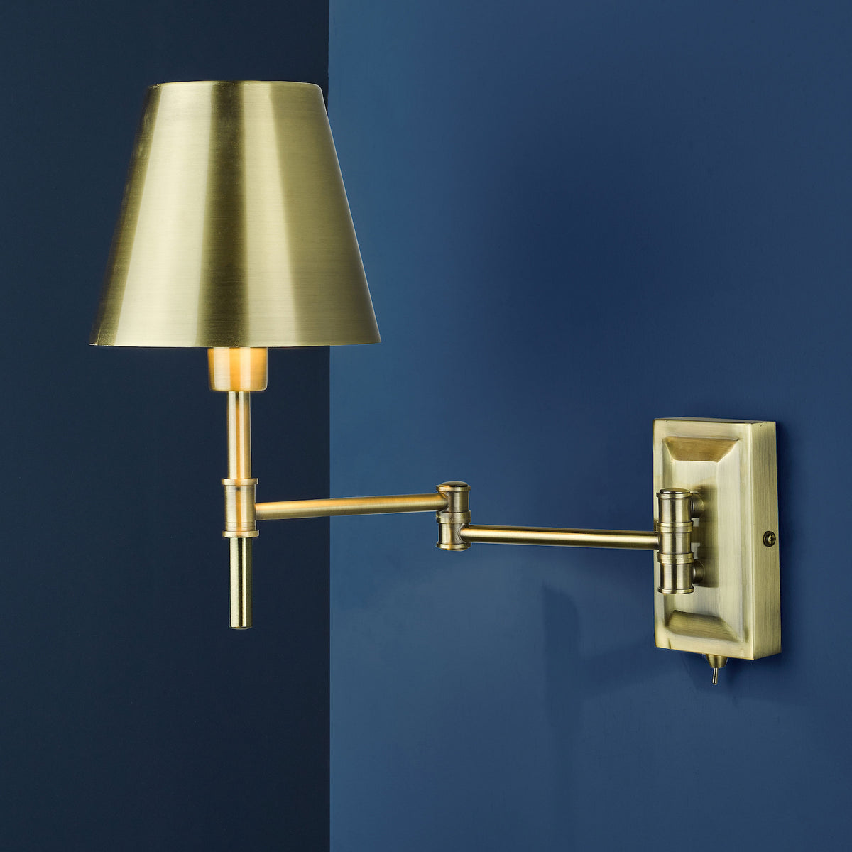 Brass swing arm wall lamp with a conical shade against a dark blue wall
