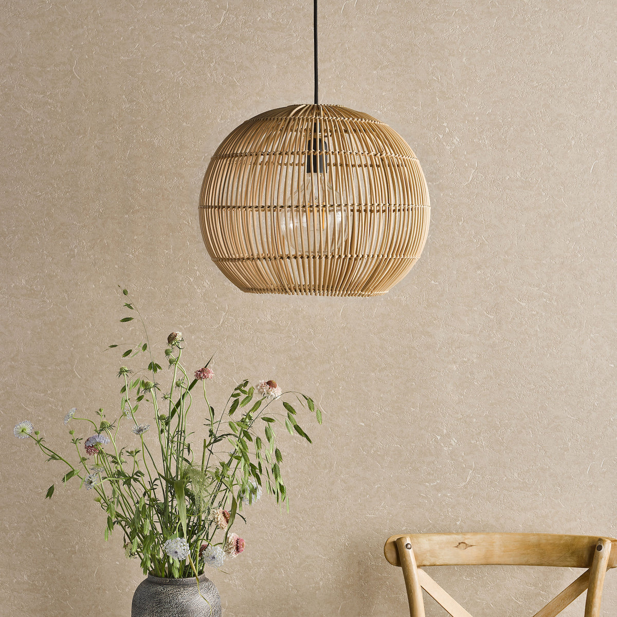 Round rattan pendant light with a clear bulb, above a vase of wildflowers and a wooden chair