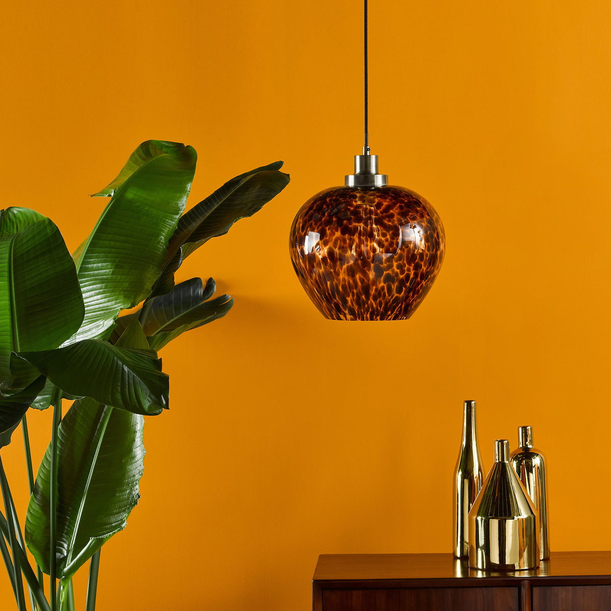 A modern pendant light with a tortoiseshell glass shade hangs above a wooden cabinet with gold vases.