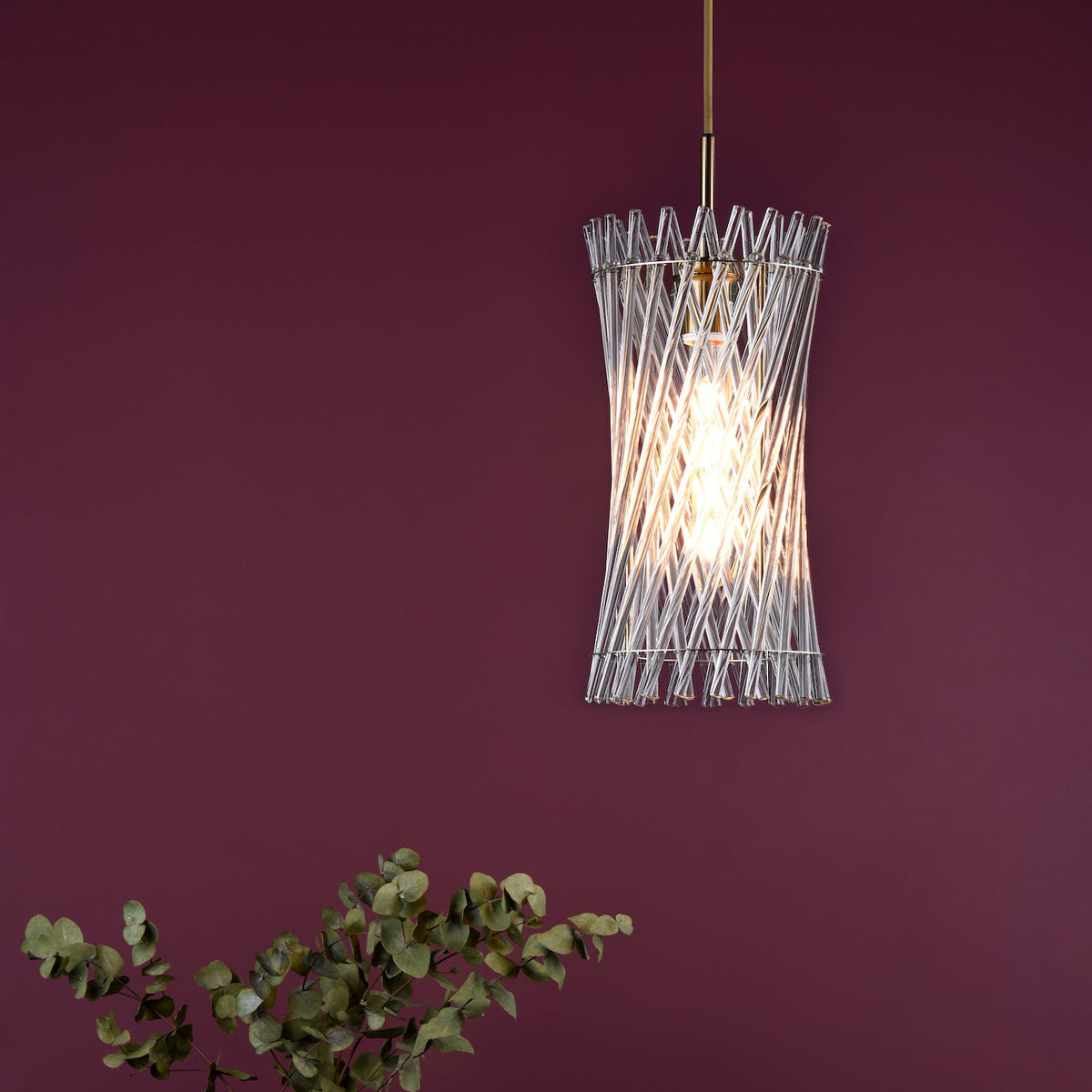 Glass rod pendant light with a gold stem and a lit bulb, against a dark red wall with green foliage