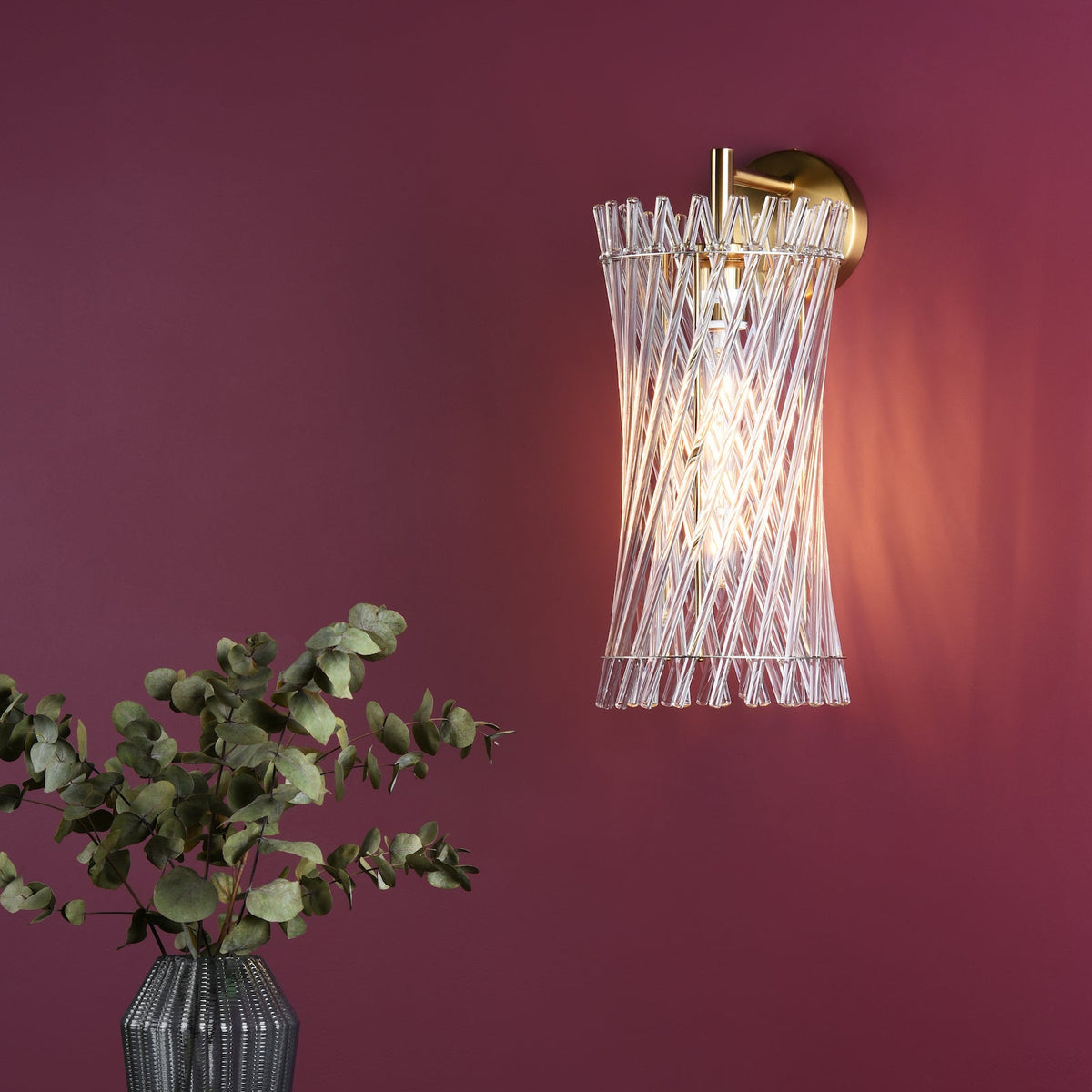 A gold wall light with a clear glass shade made of twisted rods, next to a vase of eucalyptus
