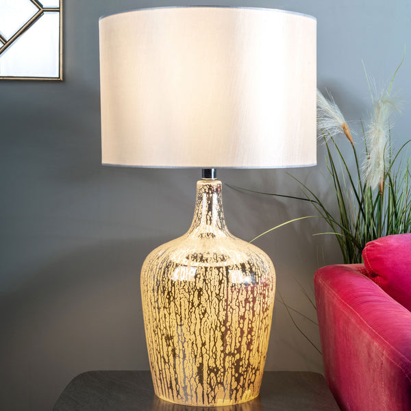 A gold mercury glass lamp with a white shade on a dark table next to a pink couch.