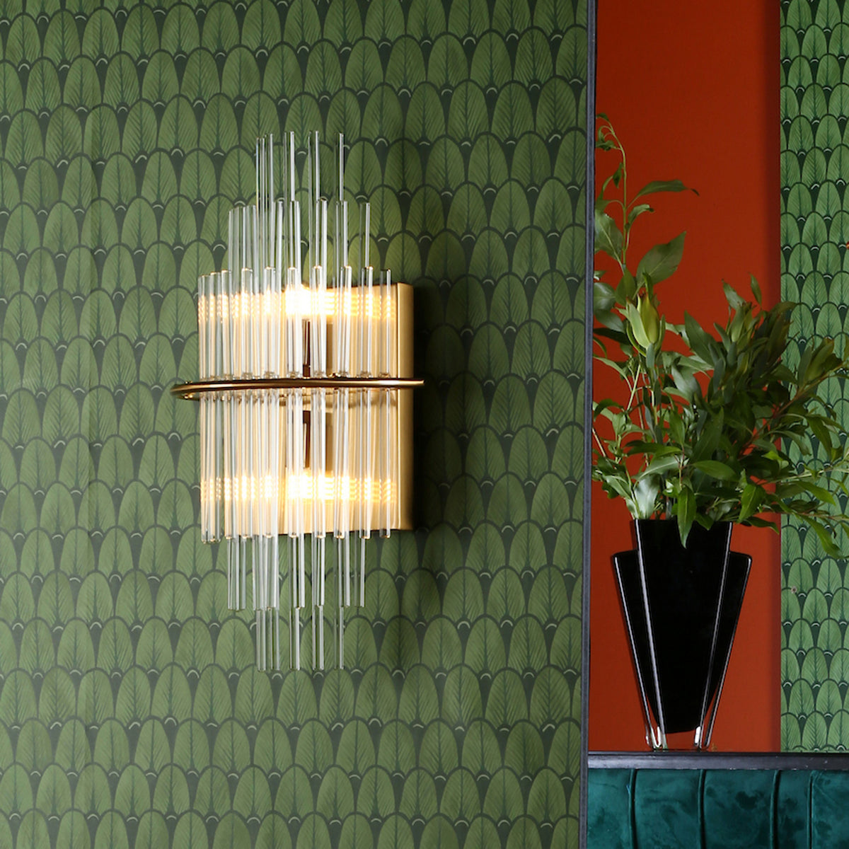 Art Deco wall sconce with glass rods and gold accents on green leaf-patterned wallpaper.