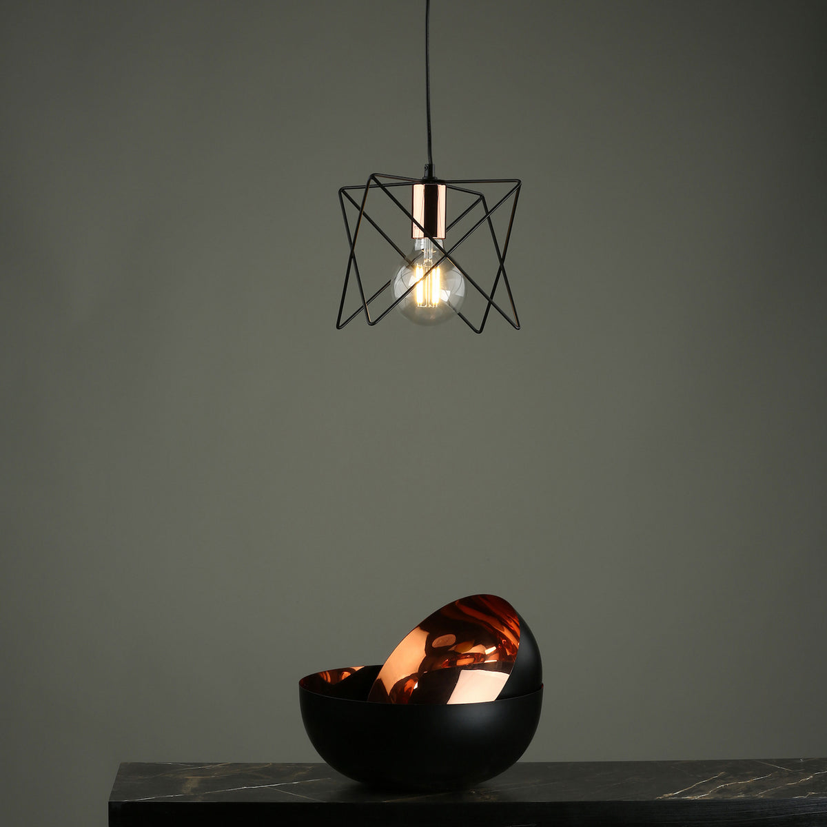 Geometric black pendant light with a glowing bulb above two black and copper bowls on a marble surface.