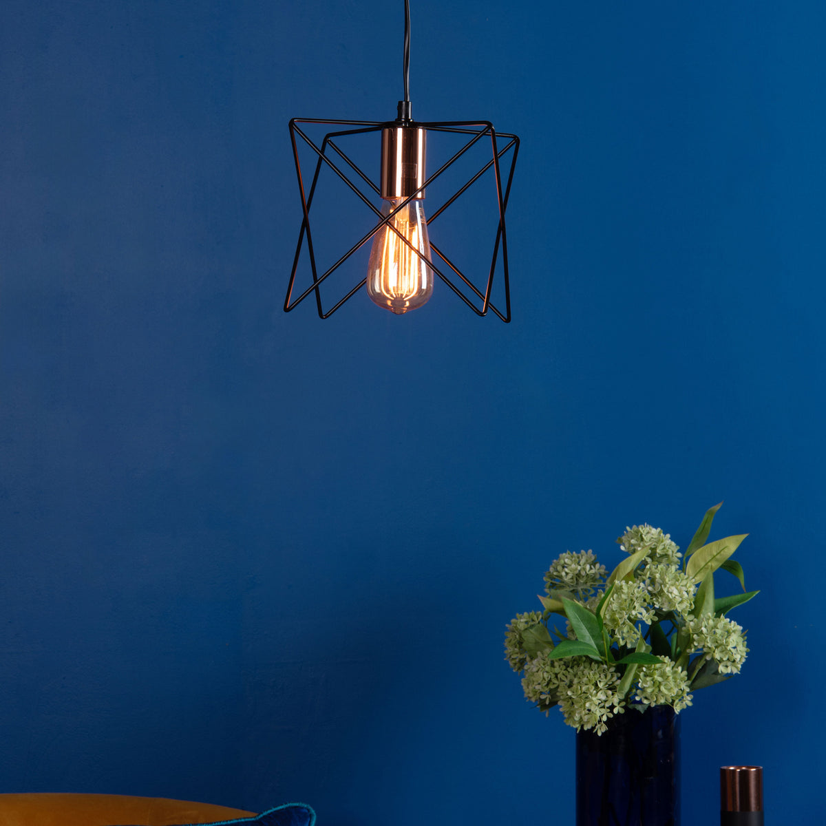 Geometric pendant light with an Edison bulb, illuminating a deep blue wall above a vase of green flowers.