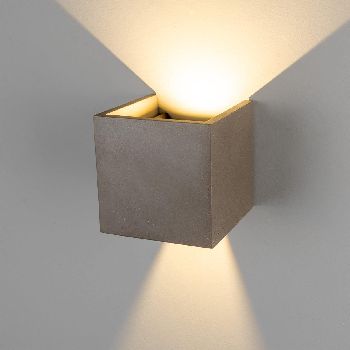 Square concrete wall sconce casting light upwards and downwards on a grey wall