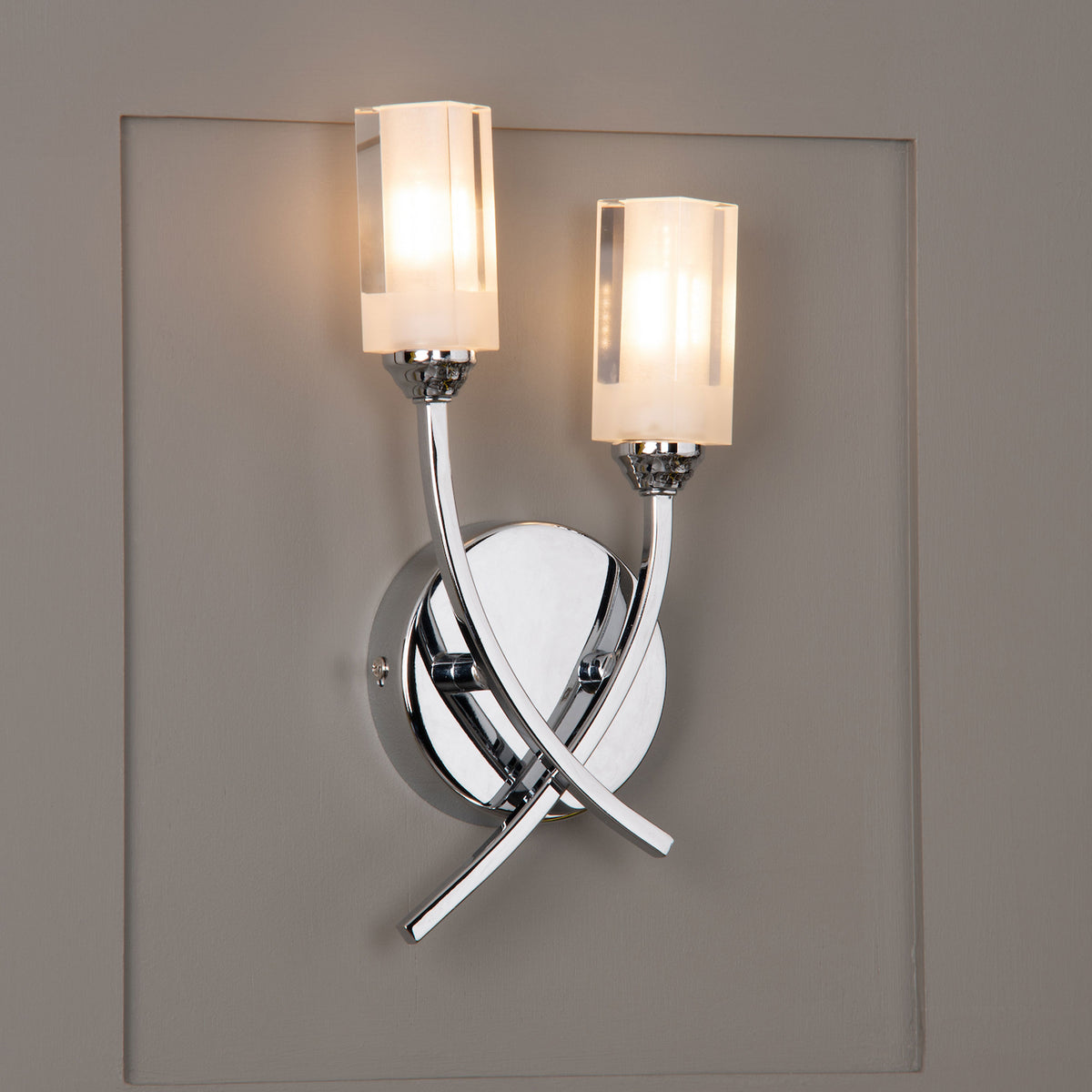 Chrome wall sconce with two frosted glass shades, casting a warm glow on a gray paneled wall. Modern design.