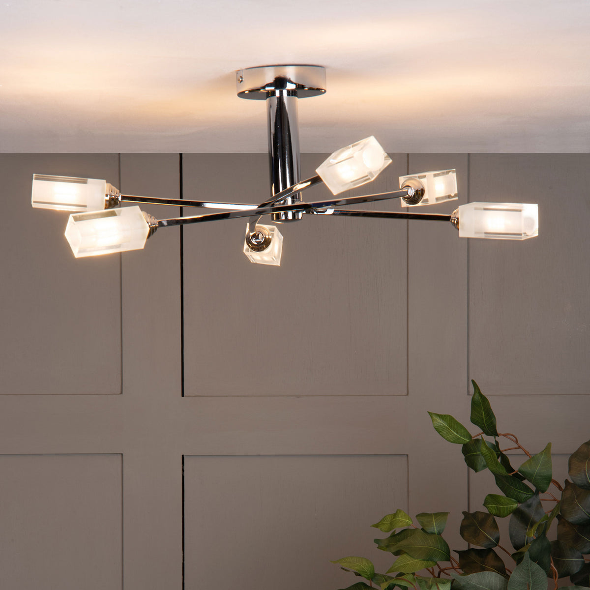 Chrome ceiling light with six frosted glass shades, illuminating a paneled wall and green plant.