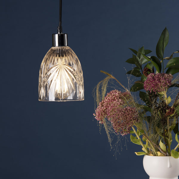 A clear, fluted glass pendant light with a chrome top, illuminating a dark blue background next to a vase of flowers.