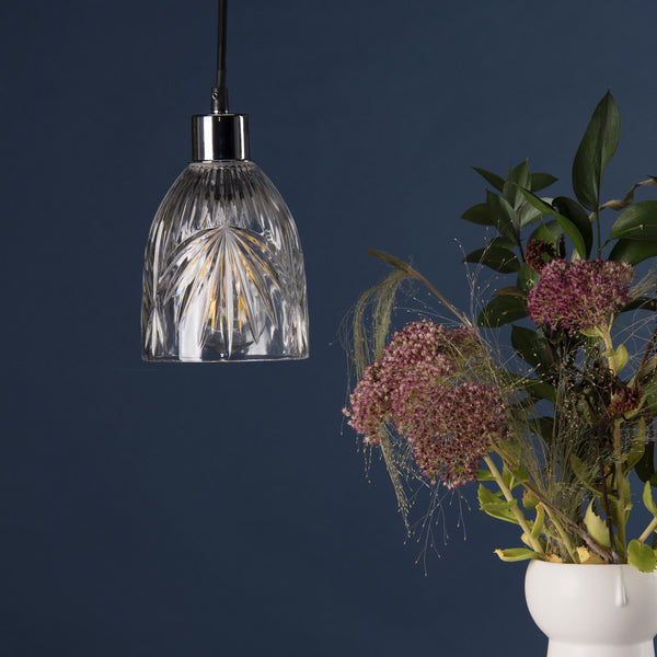A clear, cut-glass pendant light with a chrome fitting hangs next to a white vase filled with greenery and pink flowers.