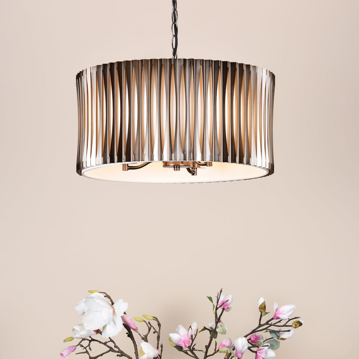 A modern chandelier with a metallic, ribbed drum shade hangs above a light pink and white floral arrangement