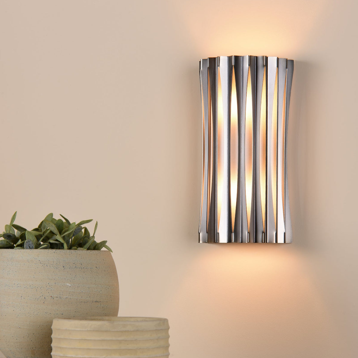 Silver wall light with vertical slats and warm glow, next to a potted plant