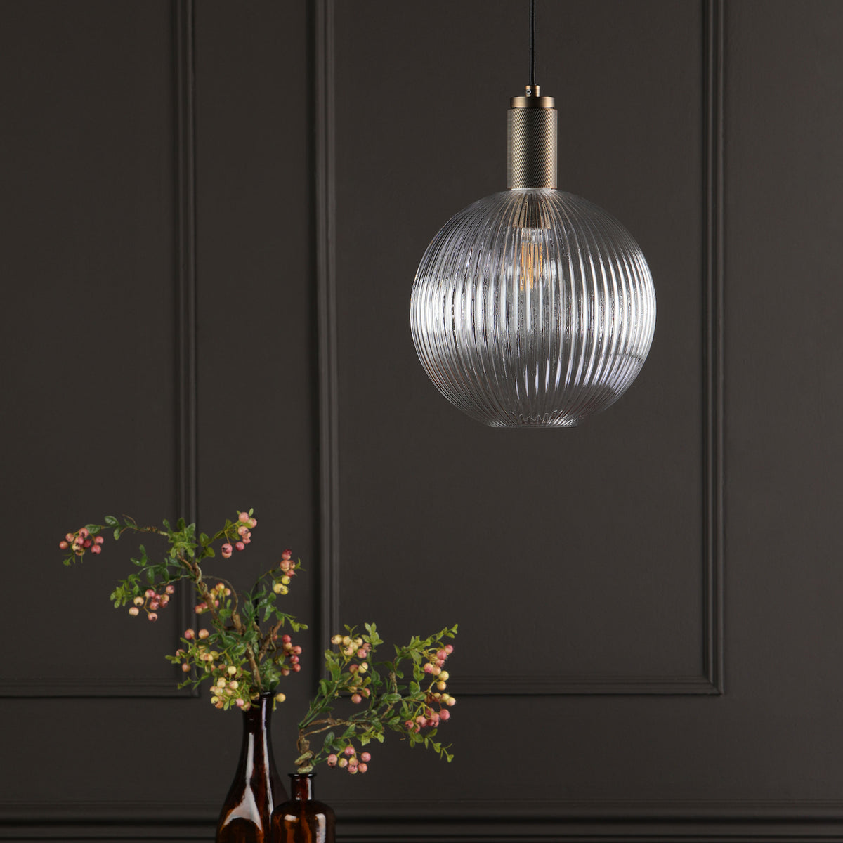 A ribbed glass pendant light with a brass top hangs above a dark brown vase with pink berries.