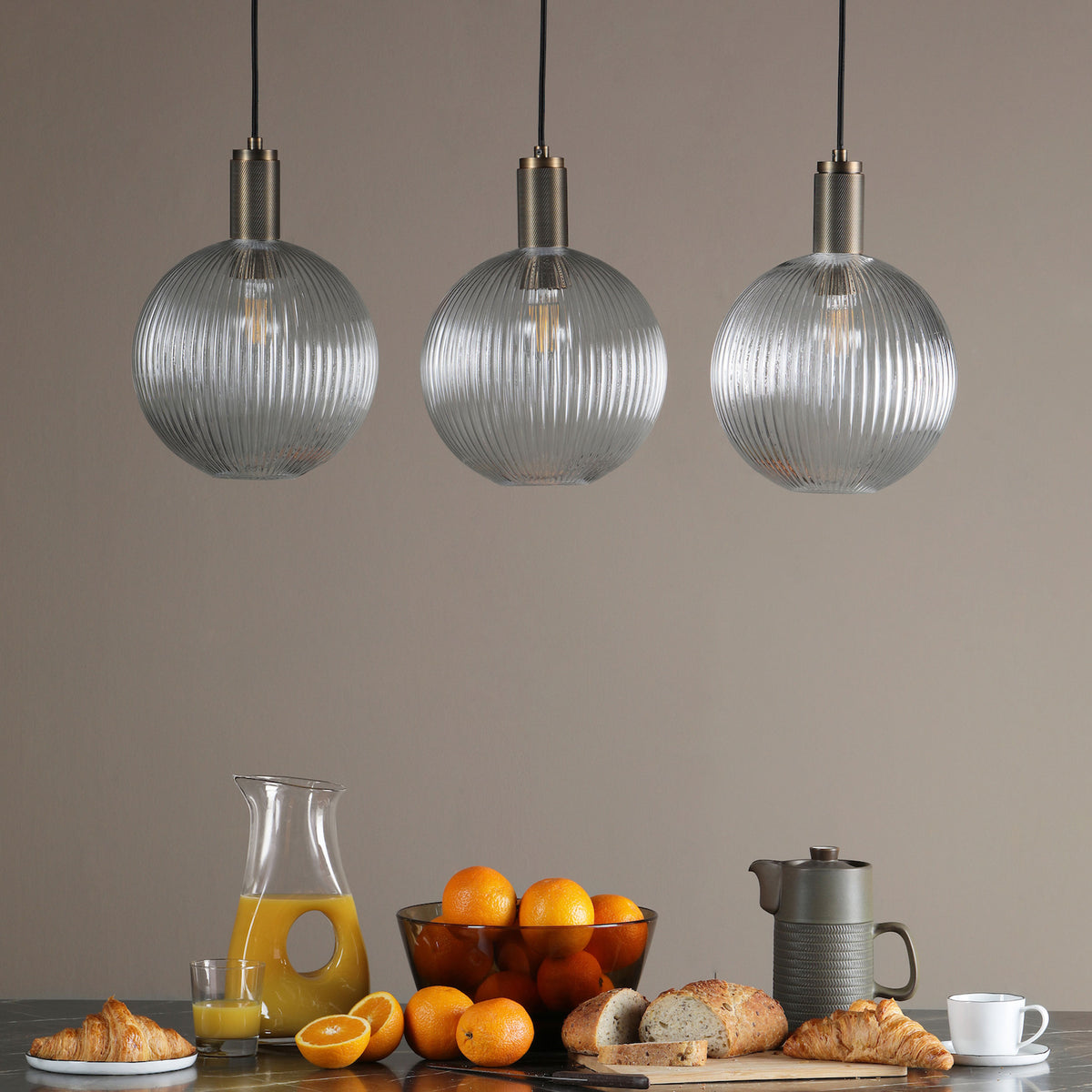 Three ribbed glass pendant lights hang above a breakfast spread of croissants, oranges, and coffee.