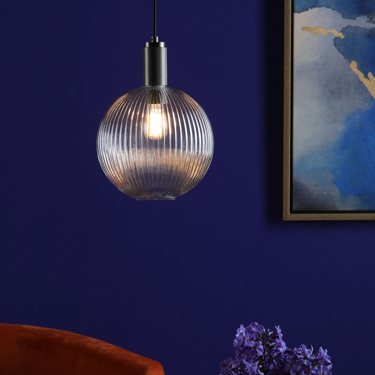 A ribbed glass pendant light with a warm glow hangs against a deep blue wall, next to a framed abstract painting.