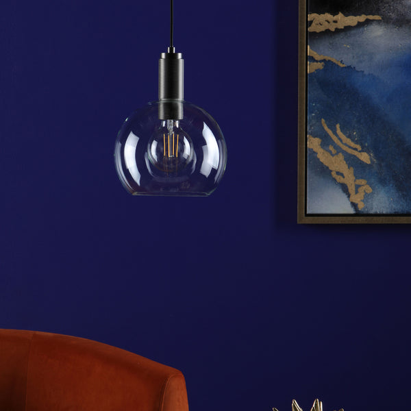 A clear glass pendant light with an exposed filament bulb hangs against a deep blue wall.