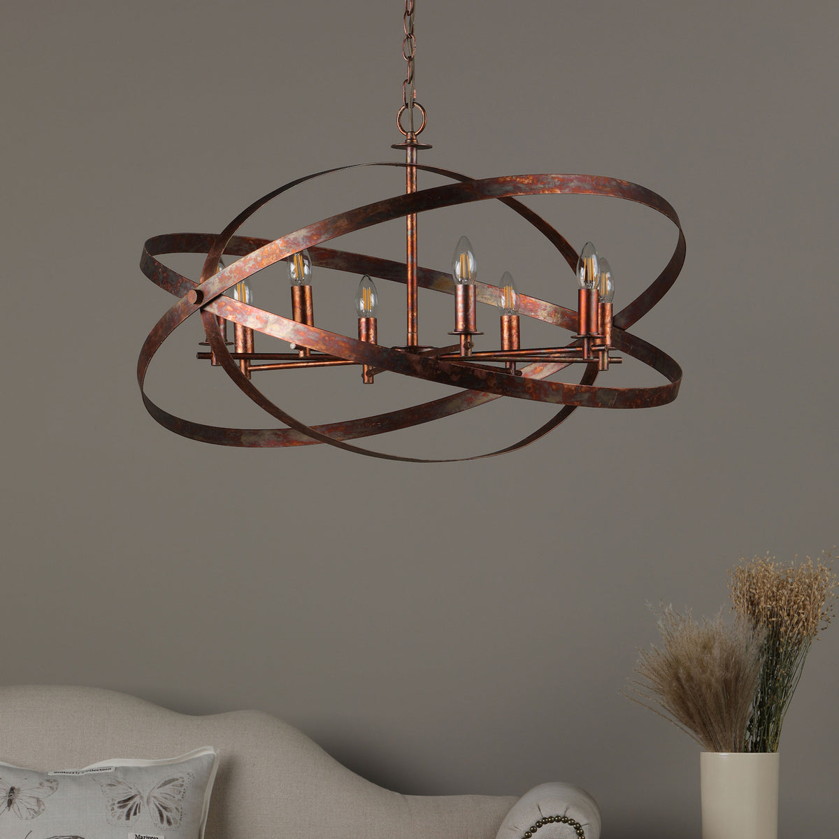 A rustic bronze chandelier with six exposed bulbs hangs above a light-colored sofa and a vase of dried flowers