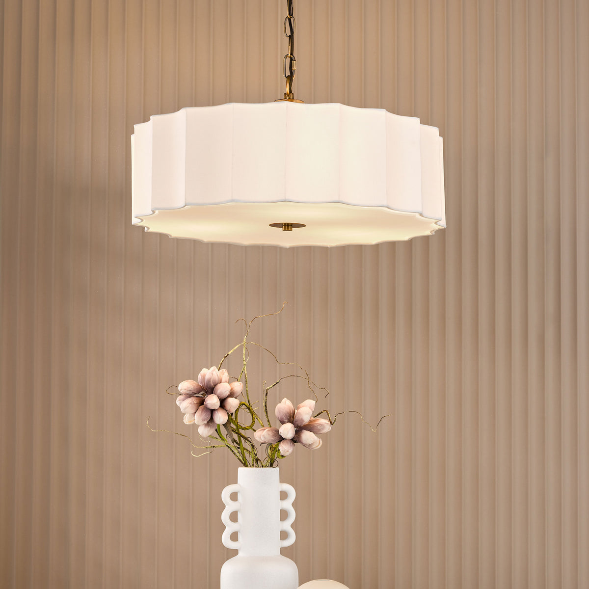 White pleated pendant light with gold chain and base, above a white vase with pink flowers and branches