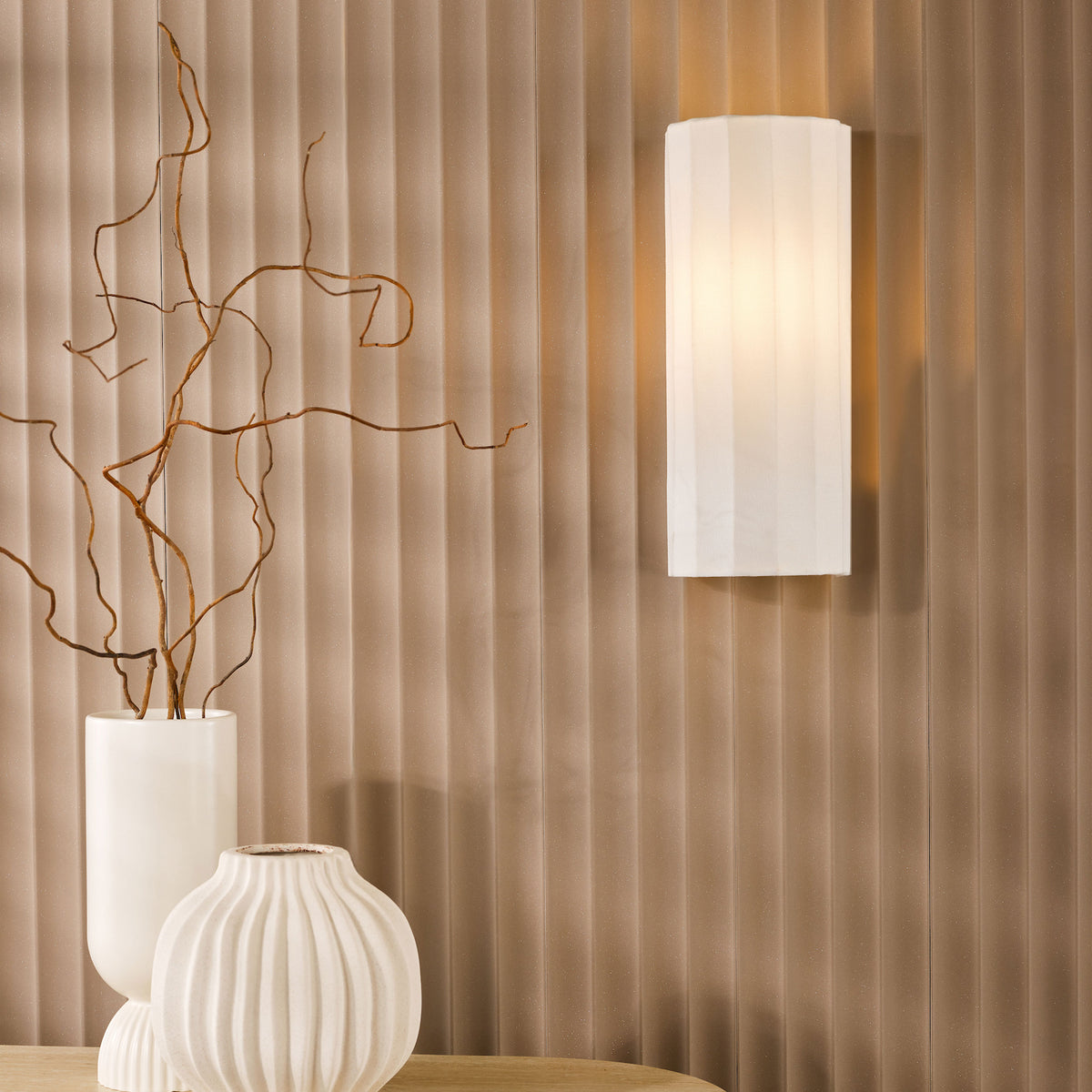 White rectangular wall sconce with light on, two white vases with branches, against a ribbed brown wall
