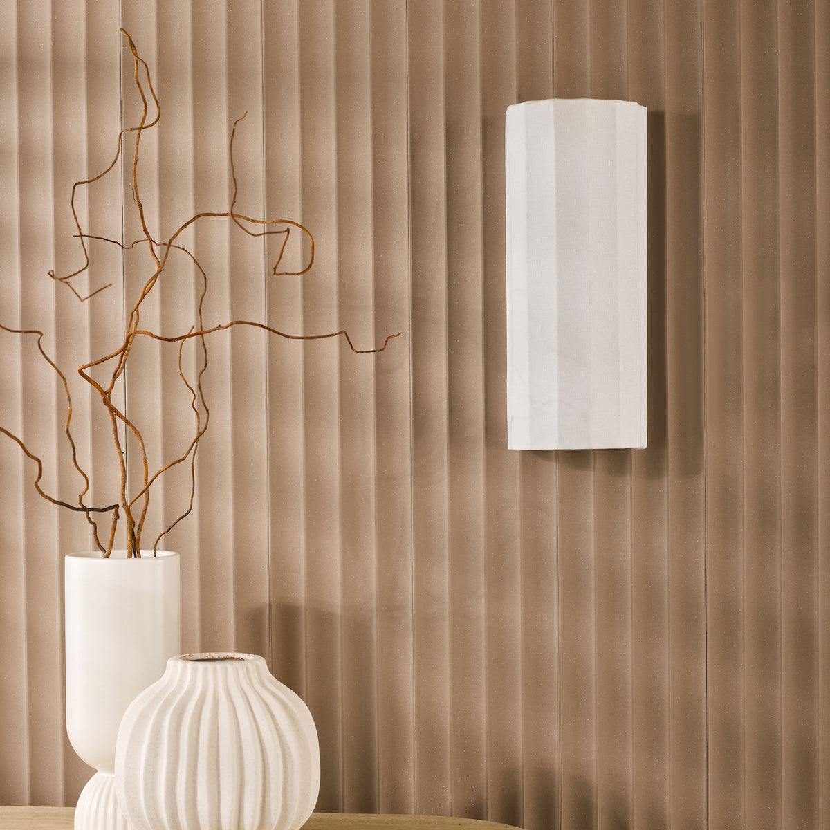 White vases with branches on a wooden surface against a brown textured wall with a white wall light