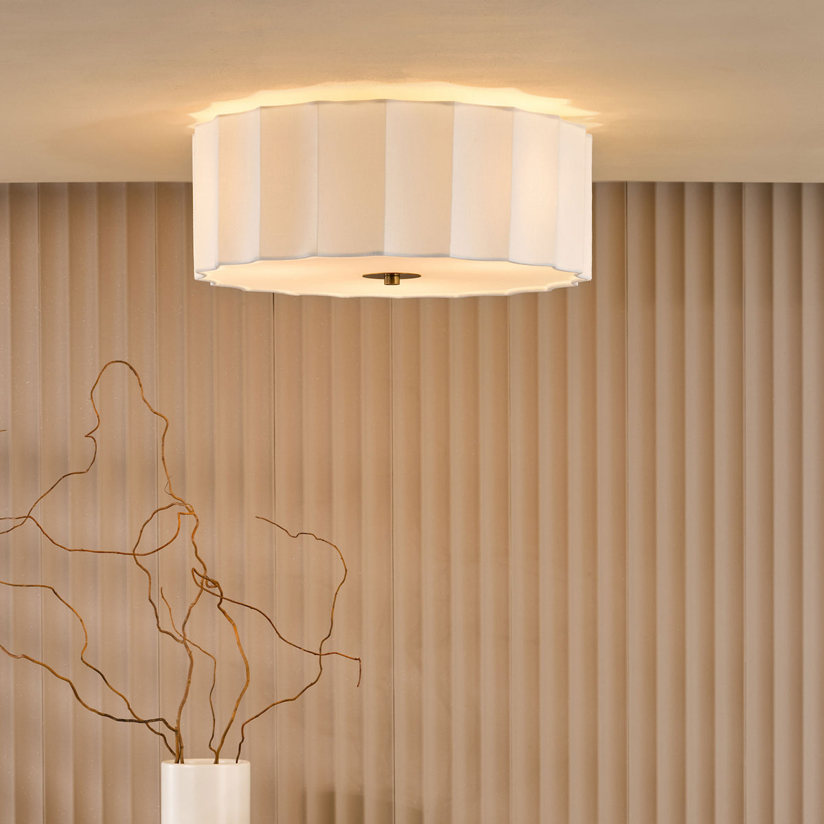 White fluted ceiling light with warm glow, above a white vase with branches, against a beige fluted wall