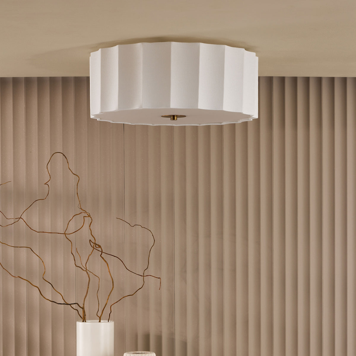 White pleated ceiling light with a gold fixture against a beige fluted wall and a white vase with branches